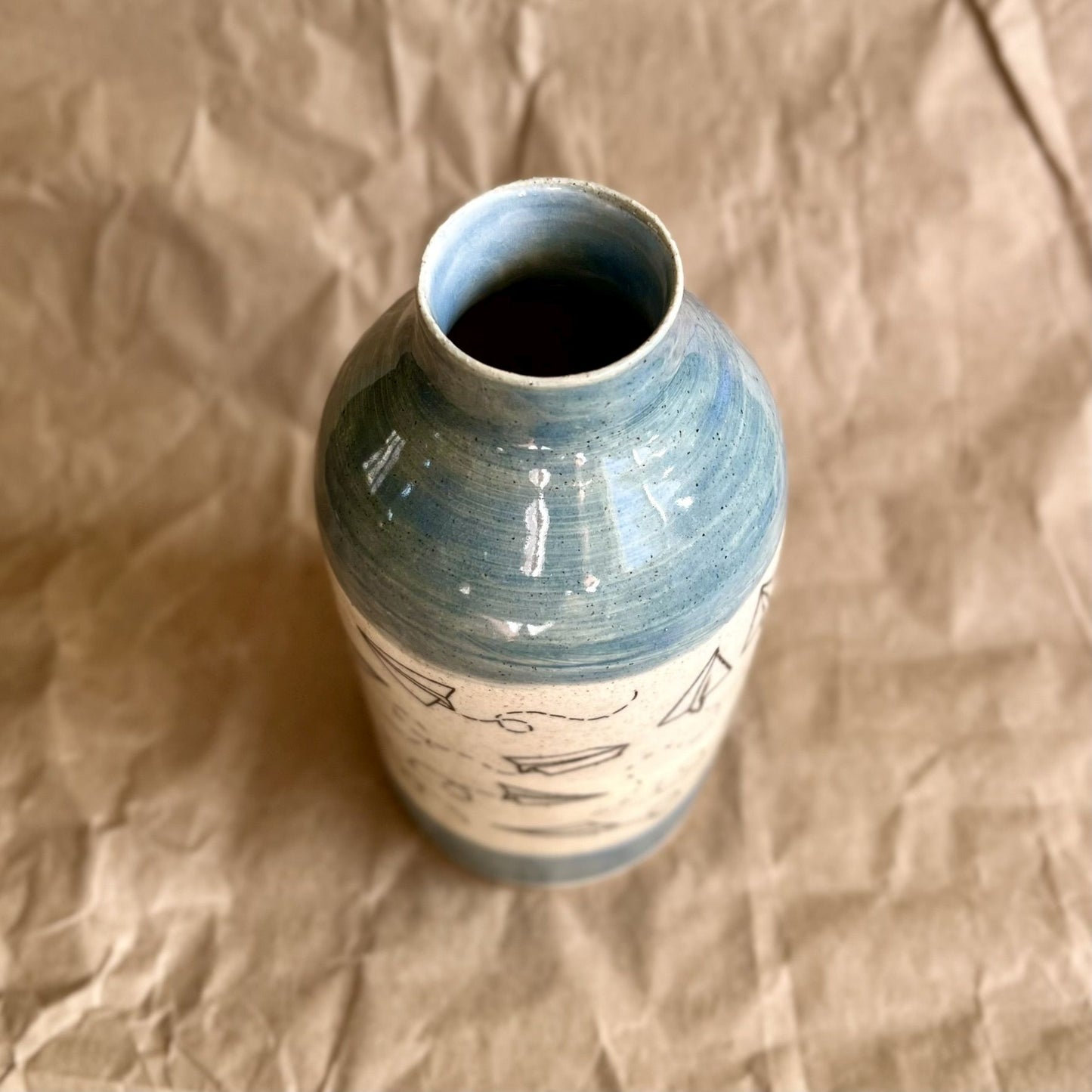 Ceramic vase with paper airplane design and blue accents on a brown textured background