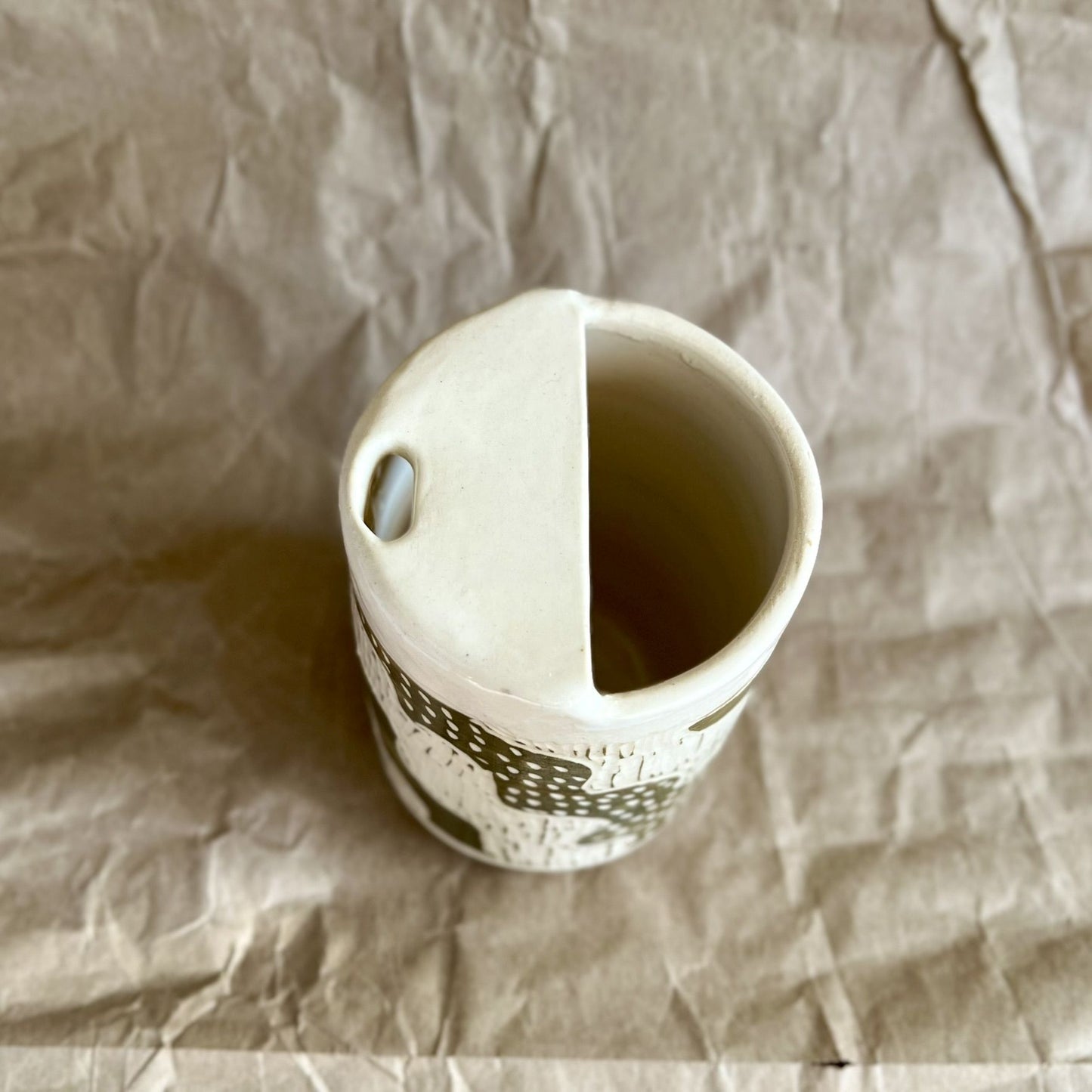 Handmade white stoneware to-go cup with green sgraffito design on a textured brown paper background