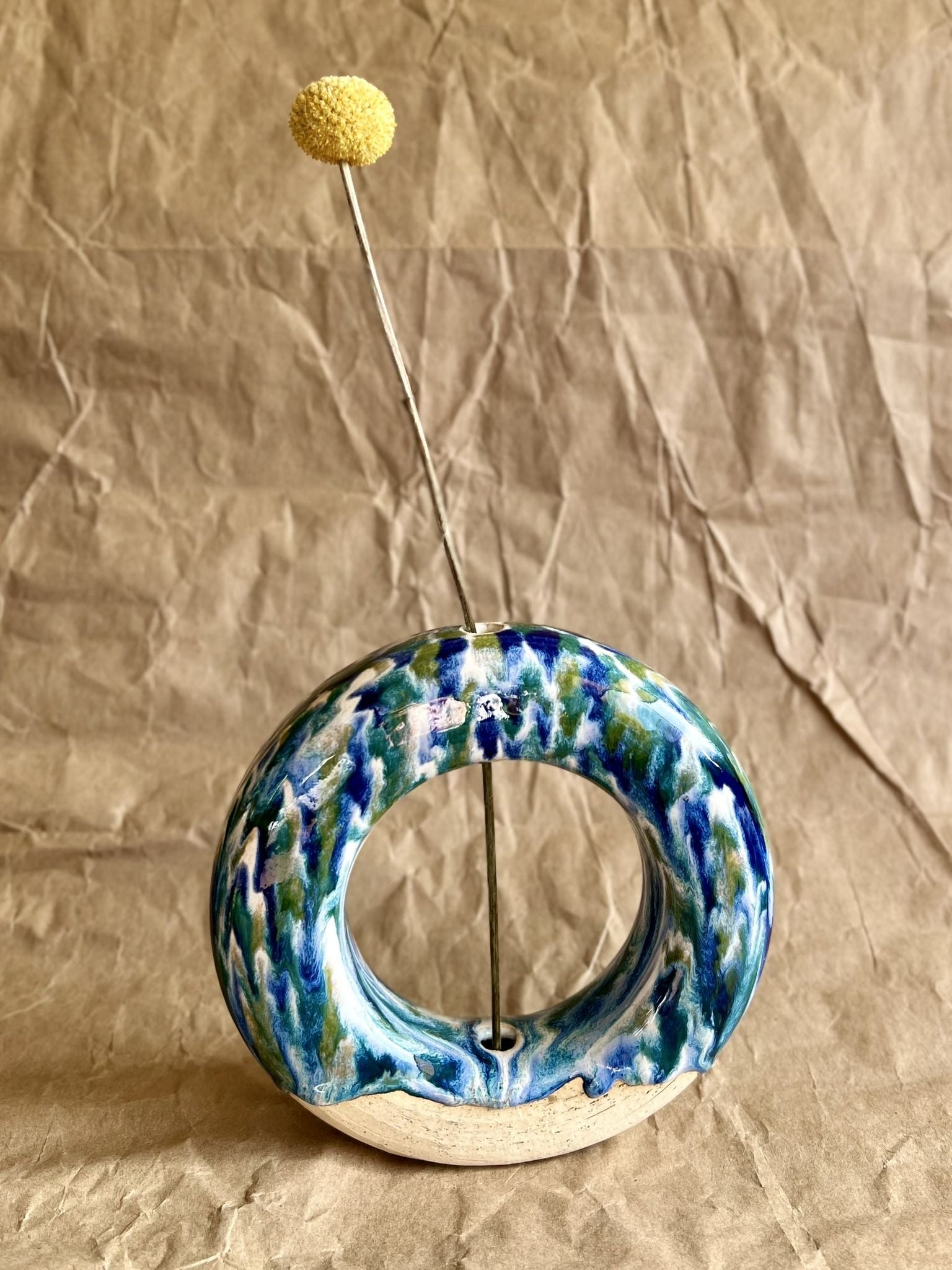 Handmade ceramic donut vase with yellow flower with blue and green glaze on a brown paper background