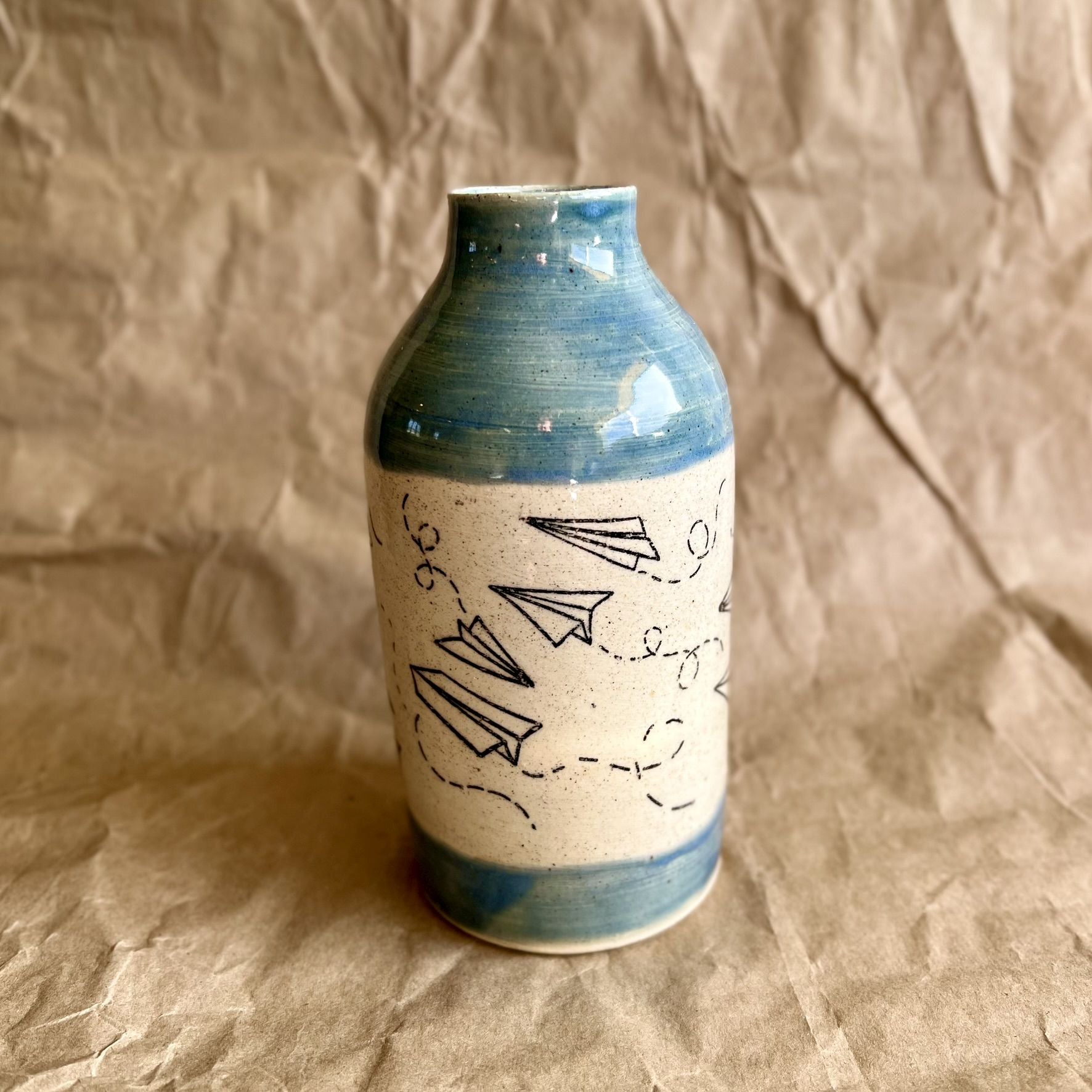 Ceramic vase with blue rim and white body featuring black paper plane line art on a brown paper background
