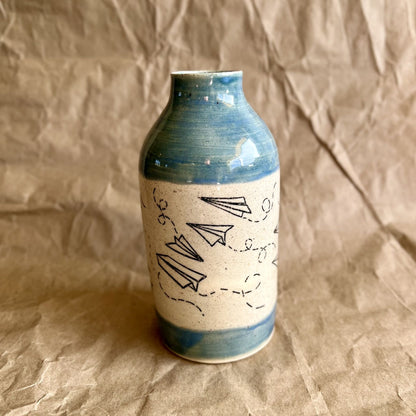 Ceramic vase with blue rim and white body featuring black paper plane line art on a brown paper background