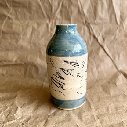 Ceramic vase with paper airplane design and blue accents on a brown textured background