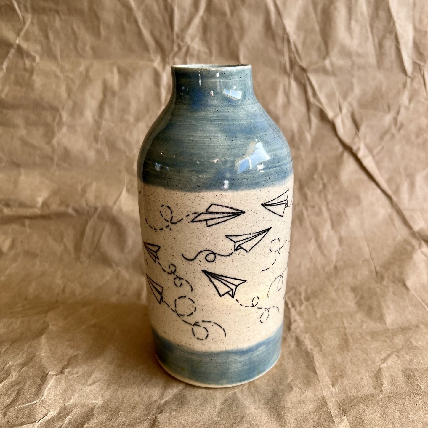 Ceramic bottle vase with blue accents and black paper plane line art on a brown paper background