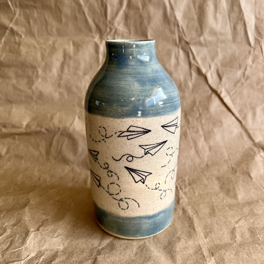 Ceramic bottle vase with blue accents and black paper plane line art on a brown paper background