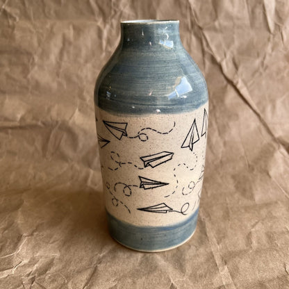 Ceramic vase with paper airplane design and blue accents on a brown textured background