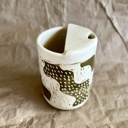 Handmade white stoneware to-go cup with green sgraffito design on a textured brown paper background
