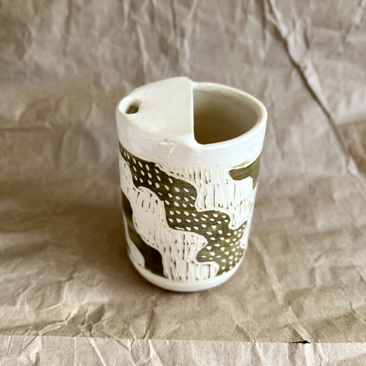 Handmade white stoneware to-go cup with green sgraffito design on a textured brown paper background