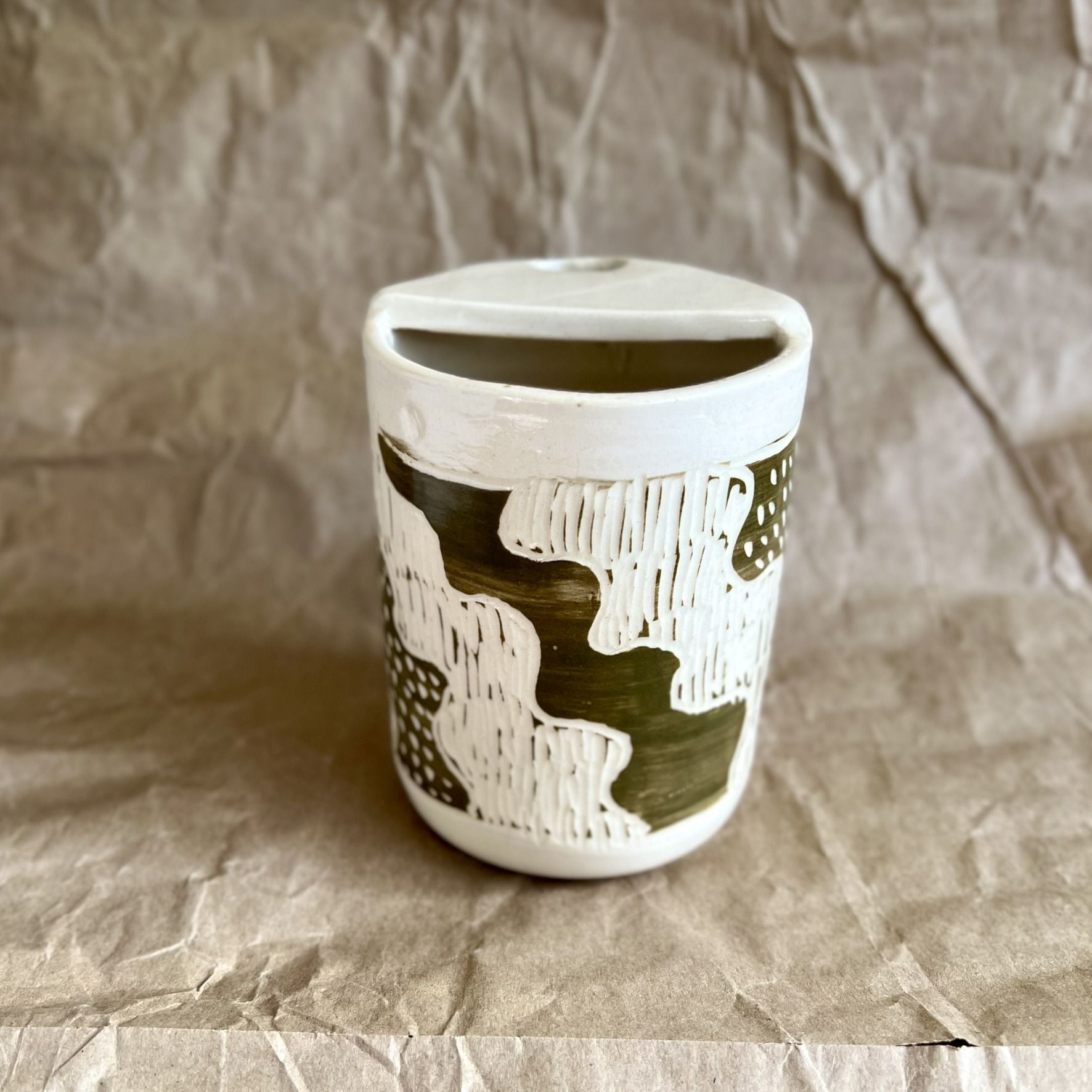 Handmade white stoneware to-go cup with green sgraffito design on a textured brown paper background