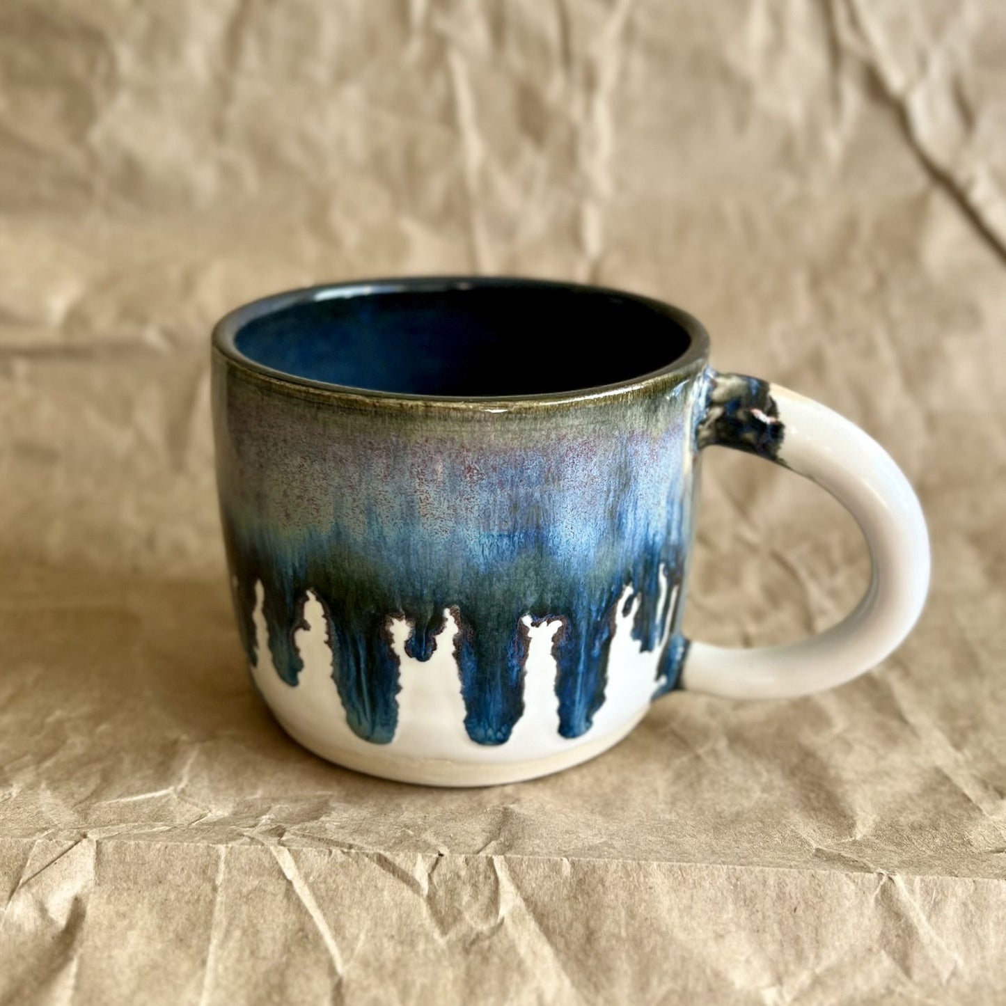 Handmade ceramic mug with blue and white drippy glaze on a crumpled brown paper surface