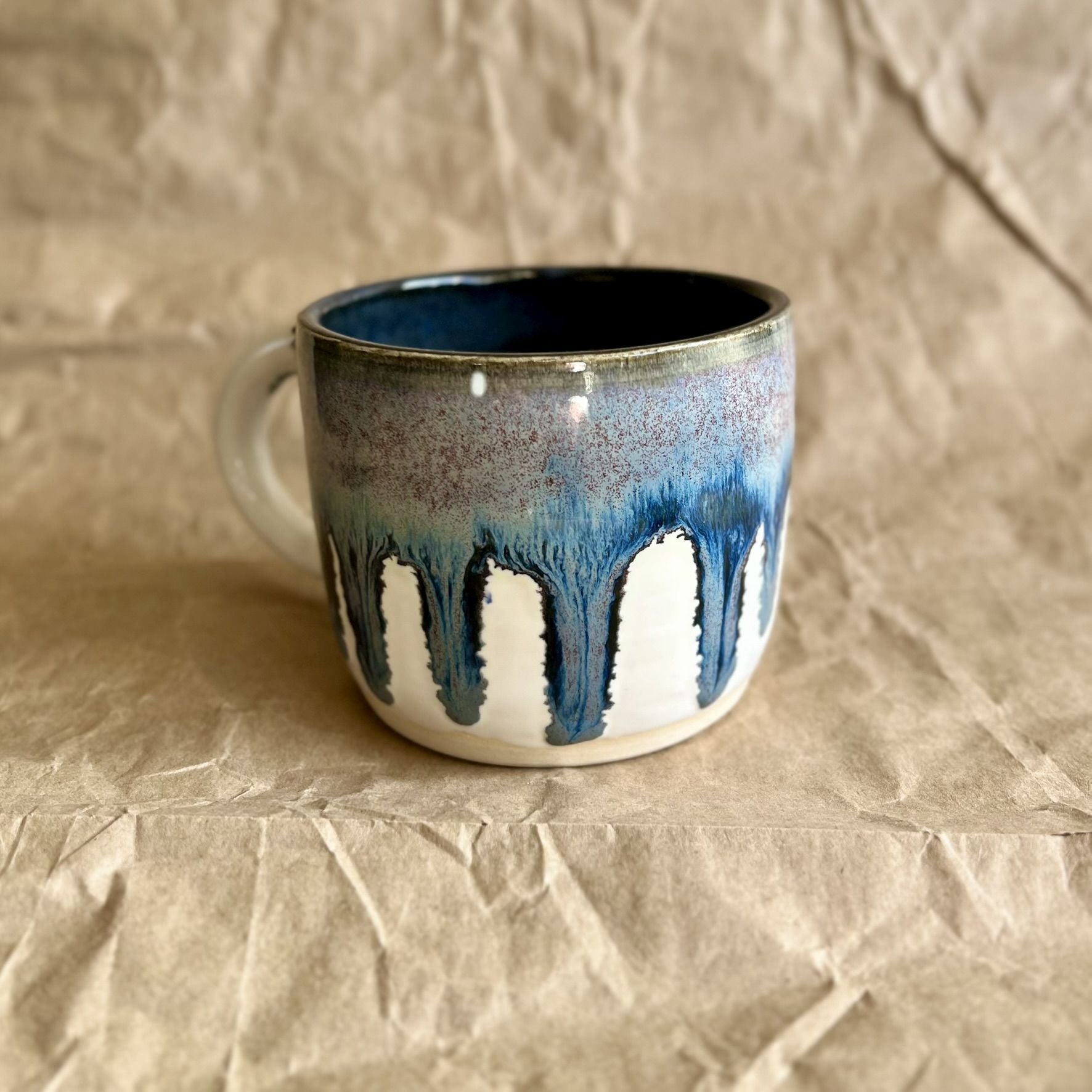 Handmade ceramic mug with blue and white drippy glaze on a crumpled brown paper surface