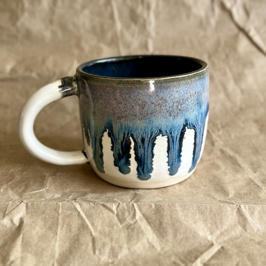 Handmade ceramic mug with blue and white drippy glaze on a crumpled brown paper surface