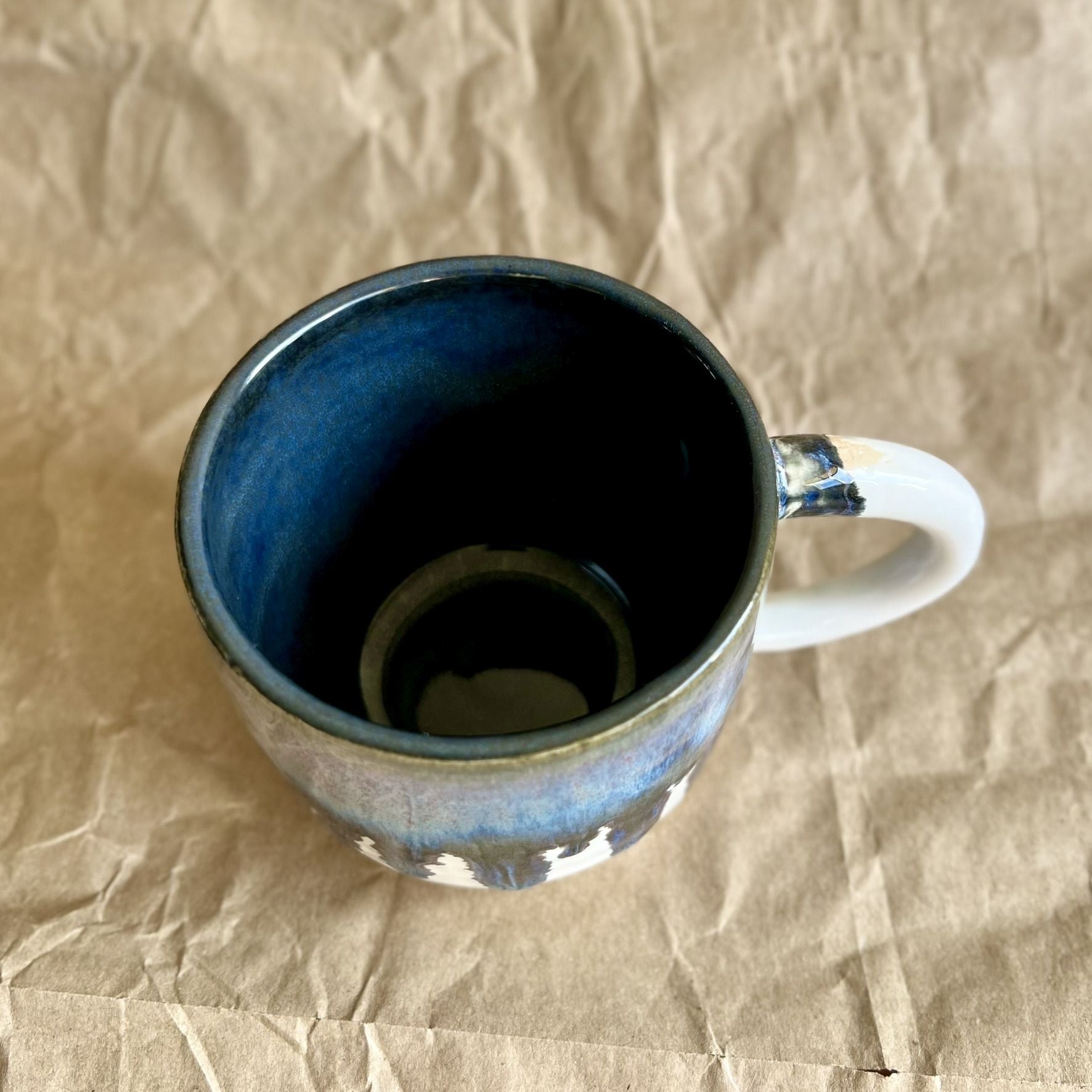 Handmade ceramic mug with blue and white drippy glaze on a crumpled brown paper surface