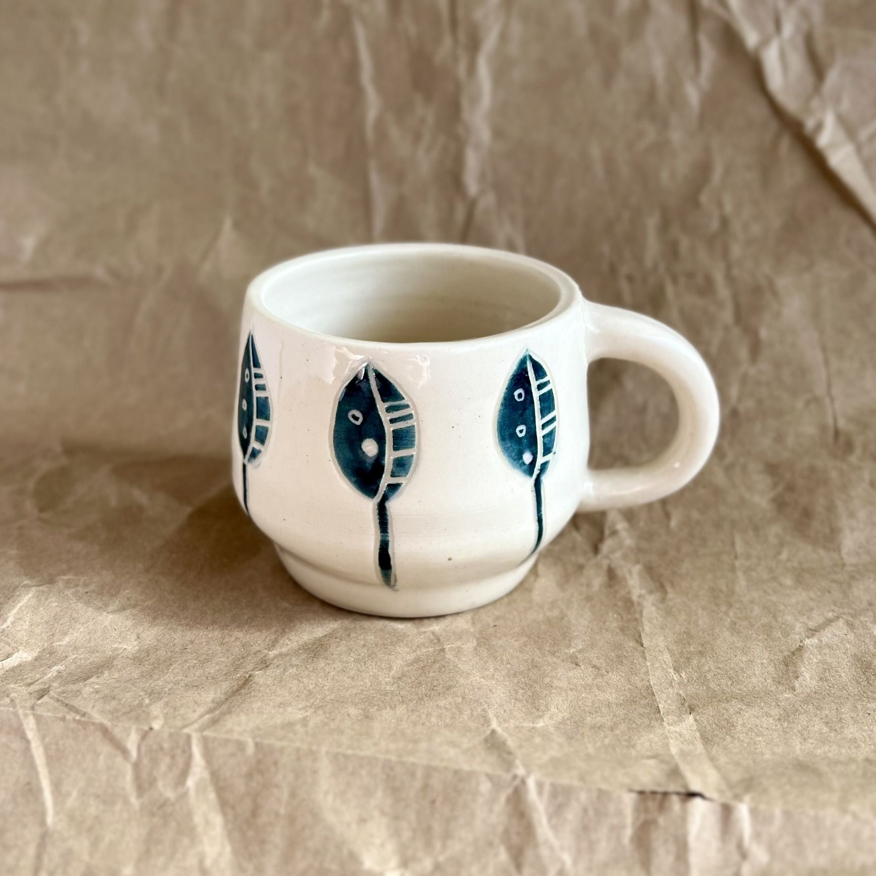Handmade white ceramic mug with blue leaf designs on a textured brown surface
