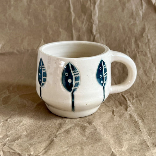 Handmade white ceramic mug with blue leaf designs on a textured brown surface