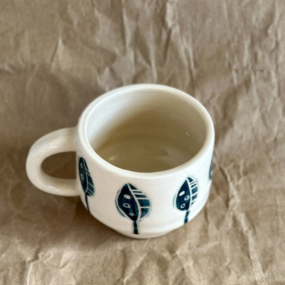 Handmade white ceramic mug with blue leaf designs on a textured brown surface