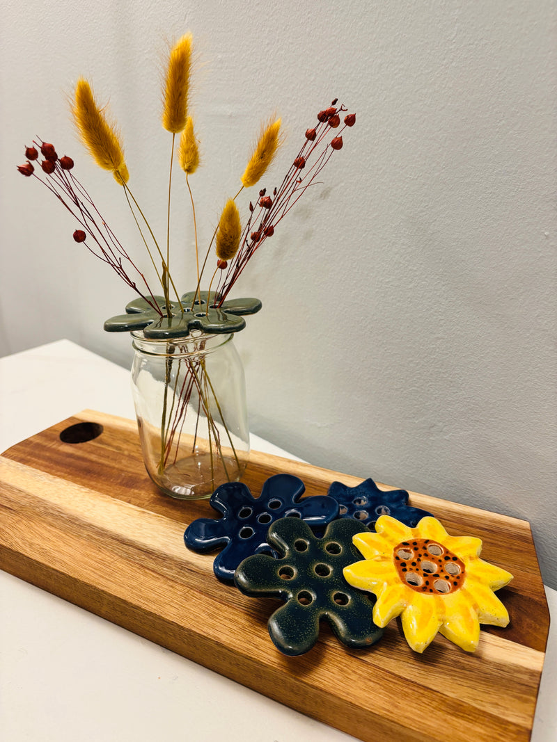 Wooden cutting board with ceramic flower frogs and a glass vase with dried plants on a white surface.