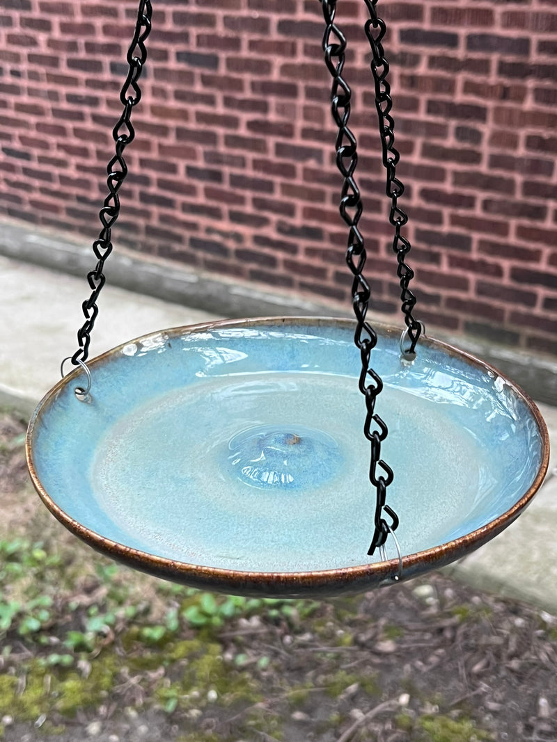 Ceramic bird bath with a blue glaze and brown rim, hanging from black chains against a brick wall.